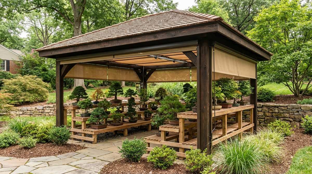 A dedicated bonsai structure with an open-front design, featuring graduated display benches arranged by tree height and a hip roof to protect specimens from excessive sun and rain. The tiered shelving accommodates various pot sizes with proper drainage, and adjustable shade screens on overhead tracks modulate summer sun intensity, casting dappled shadows to prevent leaf scorch on delicate species.