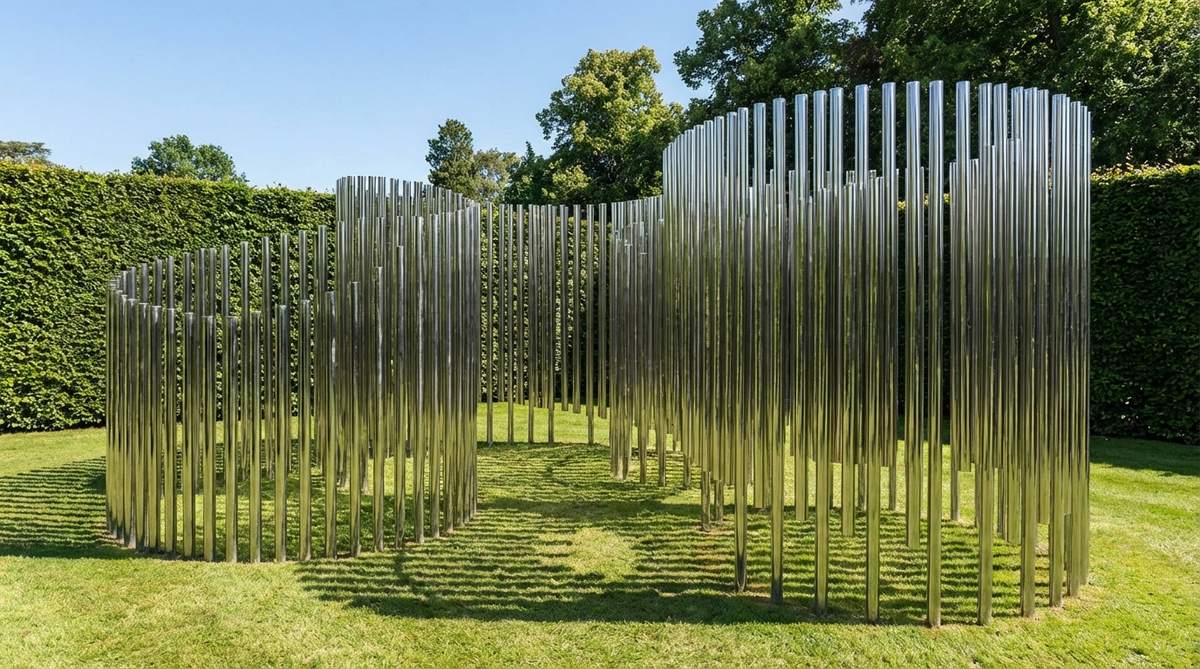 A modern garden sculpture featuring hundreds of small mirror-finish stainless steel tubes arranged vertically to create shifting, face-like forms. As viewers move, the tubes reflect fragmented, pixelated images of the environment, with light creating intricate shadow patterns. This large-scale installation serves as a focal point, best viewed against simple backgrounds like hedge walls or clear sky.