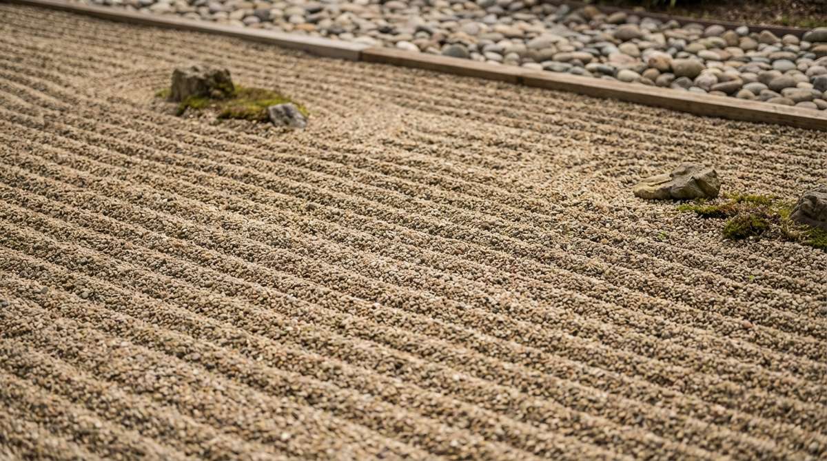 A close-up view of evenly spaced parallel lines raked into zen garden sand, representing calm water surfaces or the infinite ocean. The pattern demonstrates fundamental raking technique with consistent spacing and straight lines running the length of the gravel bed.