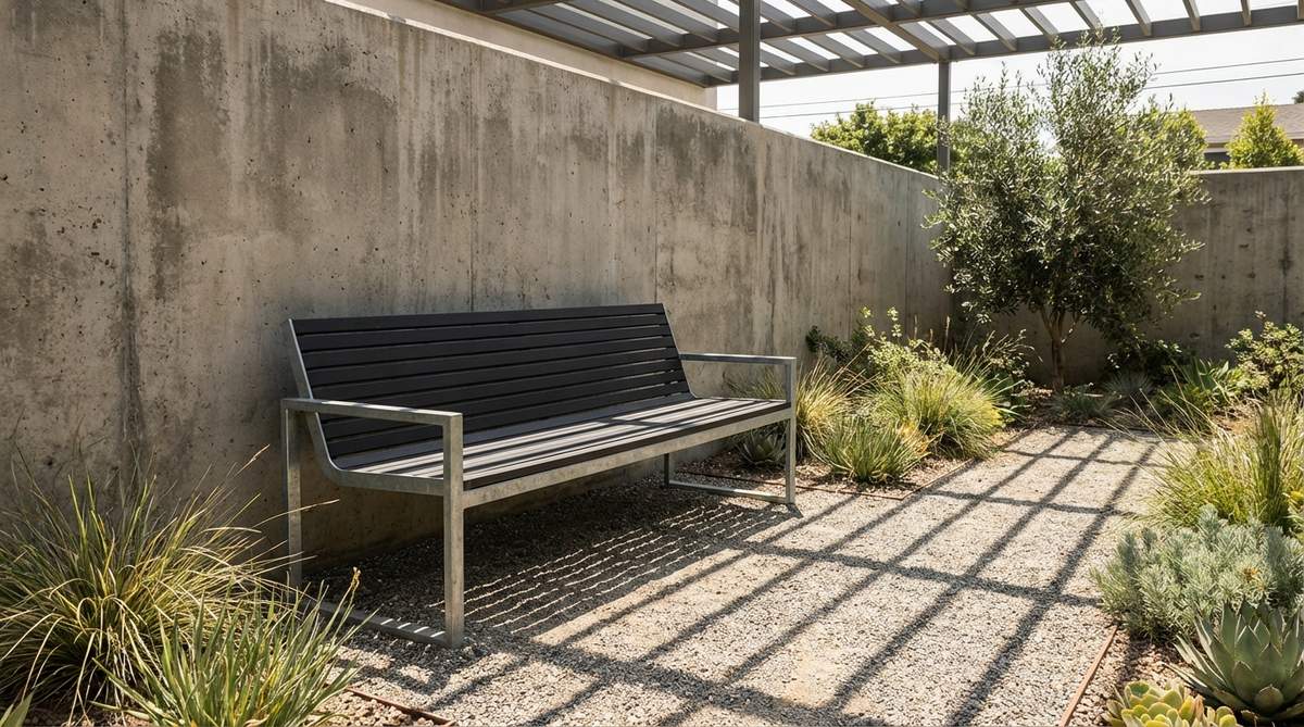 A modern powder-coated steel slat bench with galvanized frame, featuring industrial elegance suitable for urban gardens and courtyards. The bench is positioned along a wall with subtle shadow patterns from slat gaps, emphasizing geometric simplicity against greenery and gravel pathways.