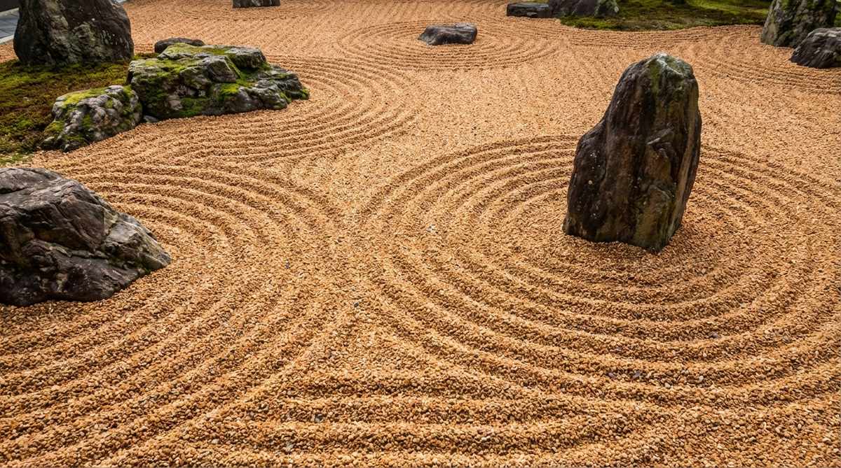 A close-up view of raked gravel in a Zen garden, depicting ocean waves with circular patterns around rock groupings, using crushed granite to symbolize water and meditative raking techniques.